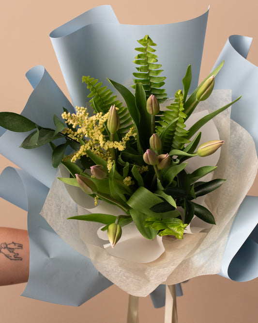 Modern floral bouquet with tulip buds, ferns, and greenery wrapped in blue paper by Annam Studio