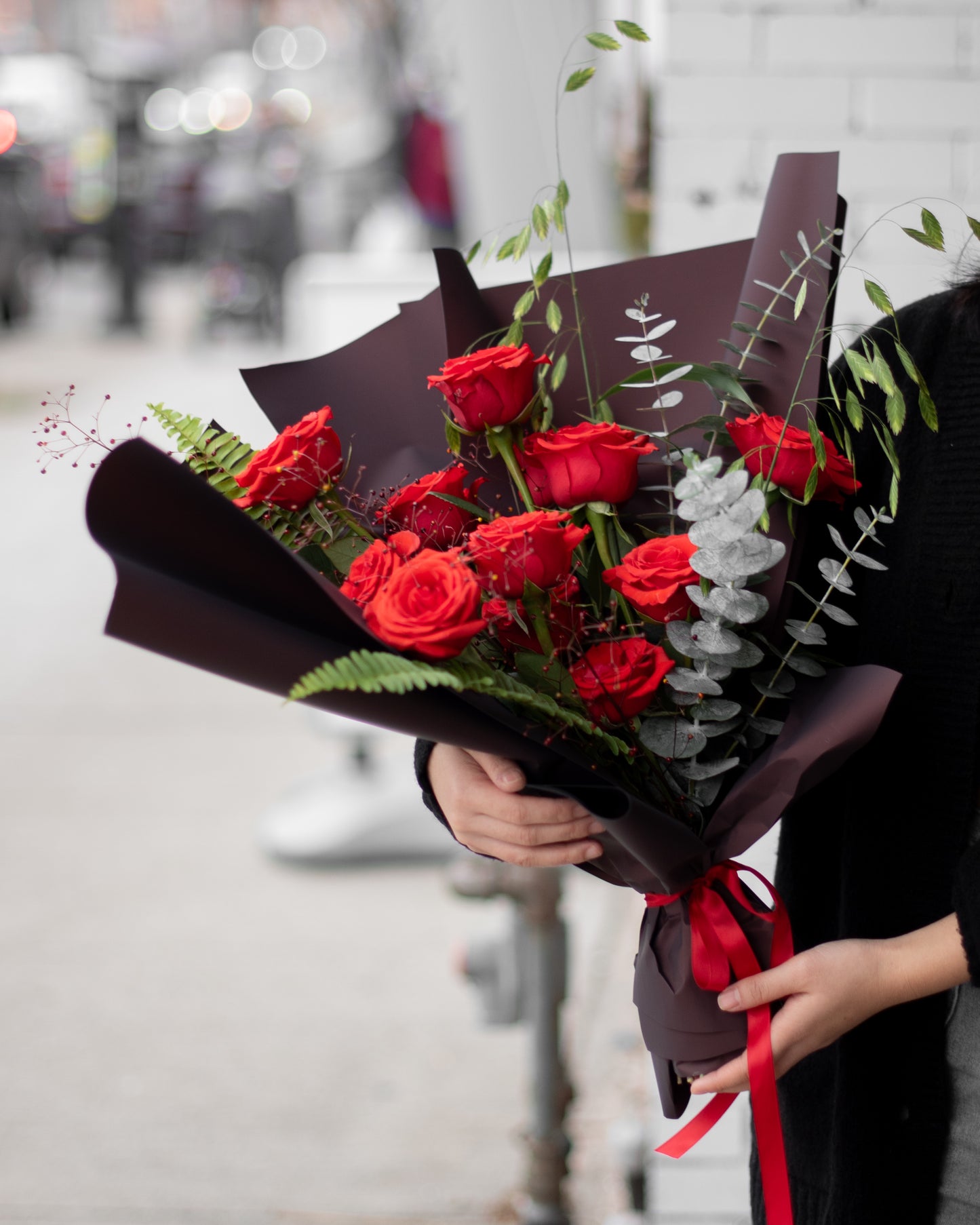 Hand holding modern red rose bouquet with greenery, wrapped in dark paper from Annam Studio Toronto